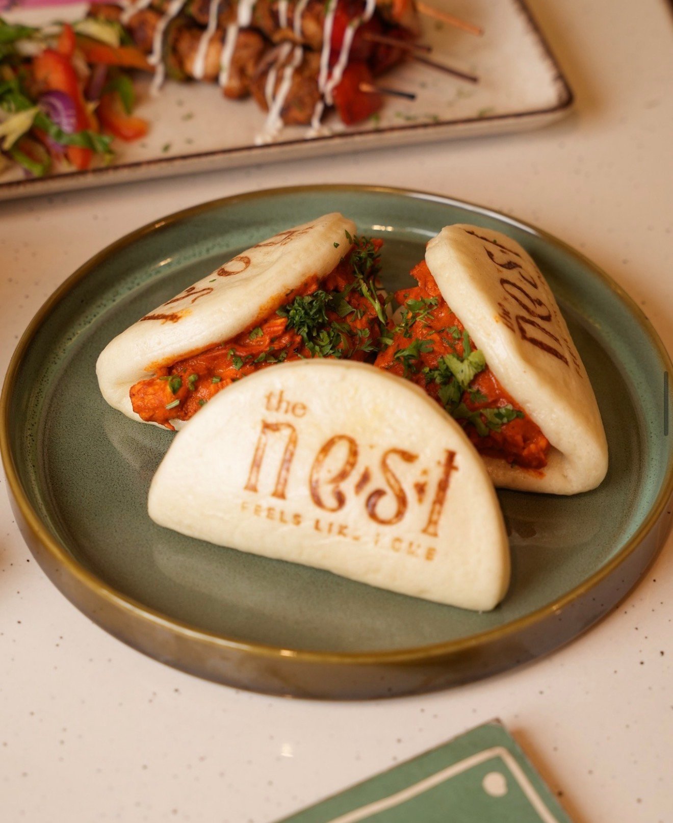 Three bao buns filled with a reddish filling and herbs on a plate, branded “the nest