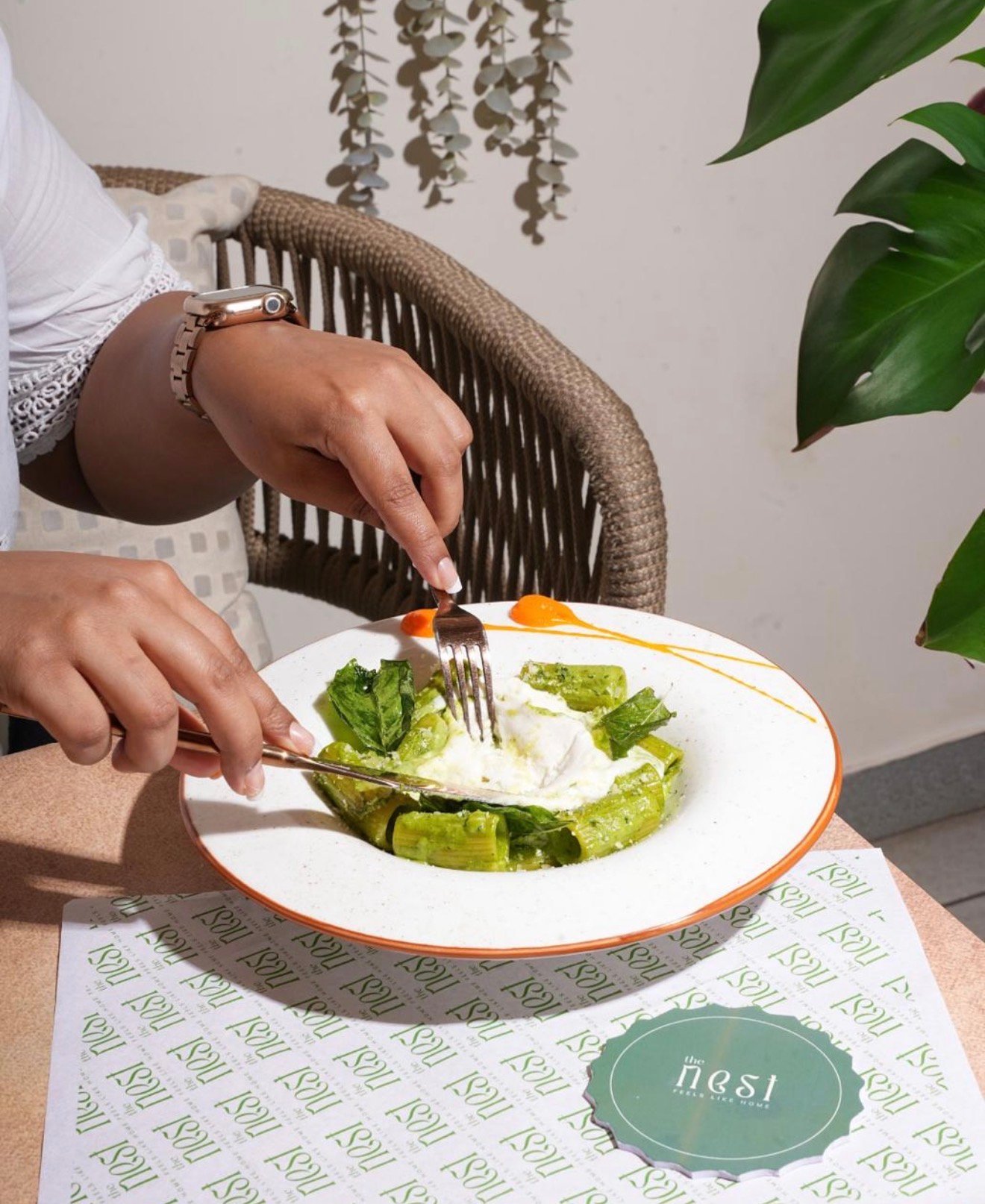 A person is cutting into a plate of green pasta with white sauce, seated at a table set with "the nest" branded paper.